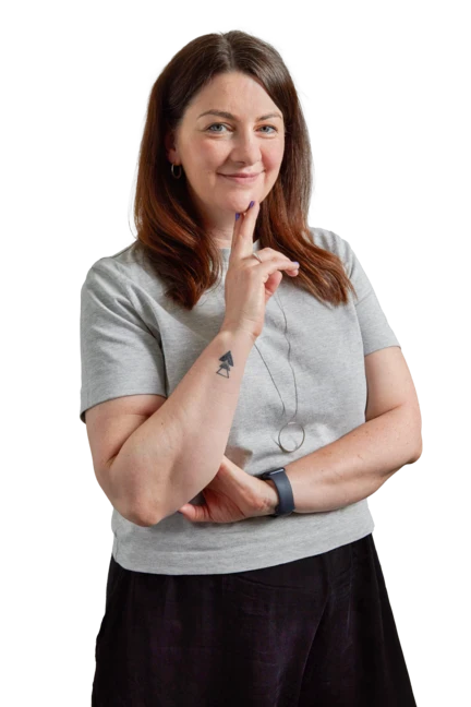 Siobhan is wearing denim jeans and a black cardigan as she leans on a wooden stool with one hand and looks directly into the camera, smiling.